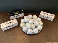 Photo showing 3 boxes of new Dunlop DDH golf balls and 19 additional used golf balls placed in a shallow bowl