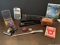 Desk with black vintage Swingline stapler, white and gray calculator, small wooden box, playing cards, paper clips, binder clips, letter opener, pencil sharpeners, clamp, and blank writing notes.