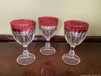Three vintage goblets with cranberry-colored flash trim around the rim, clear faceted glass stems and bowls, arranged side by side on a brown surface.