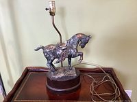 Front view of a vintage metal horse lamp showing the horse sculpture on a wooden base with a bulb socket on a curved rod and visible electrical cord.