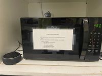 Front view of black Amazon Basics microwave oven with closed door, digital display showing 4:30, keypad control panel visible, placed on a white shelf with a small device beside it.