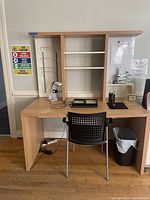 Desk 1 showing light wood desk with built-in central shelving in white, attached side metal racks, whiteboard on side panel, black plastic chair, and small trash bin below.