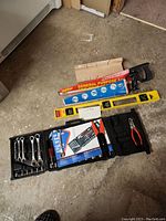 Photo of assortment of tools showing 2 saws, a general purpose saw blade in packaging, a yellow spirit level, and a black tool kit containing wrenches, screwdriver bits, and pliers on the floor.