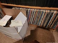 Open cardboard box with vinyl records in white and brown sleeves beside wooden shelf filled with vinyl records