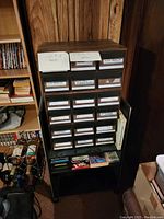 Photo of black wooden cassette tape organizer with multiple labeled drawers and cassette tapes on bottom shelf, showing overall lot contents.