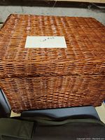 Photo of a large rectangular wicker basket with a lid placed on a surface against a wall. The basket is woven from natural fibers and has a uniform weave pattern.