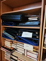 Shelf with multiple black, blue, and other colored binders stacked horizontally and vertically. White cards label 'Lot #14 binders' and 'Lot #10 jewelers' are visible.