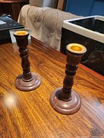 Photo showing pair of vintage wooden candle stick holders on a wooden table with a dark trash bin and beige fabric in the background.