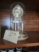 Decorative anniversary clock inside a glass dome on a carved base, sitting on wooden shelf with a paper label reading 'Lot #42 Clock'.