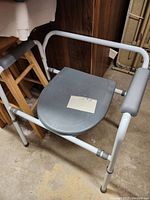 Portable commode chair with gray plastic seat and lid, white tubular metal frame, and dark gray padded armrests, shown from a slightly angled front.