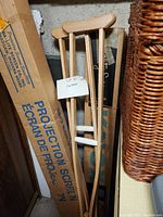 Two wooden adjustable crutches leaning against boxes, equipped with padded hand grips and adjustable metal pegs for height adjustment.