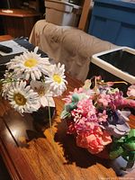 Photo of two faux floral arrangements placed on a wooden table, one with white daisies and the other with mixed pink and purple flowers.