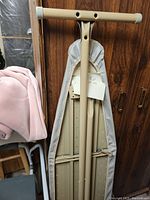 Full view of the ironing board with beige fabric cover and metal frame leaning against a wooden door.