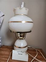White vintage glass lamp with gold trim placed on wooden surface with power cord; includes handwritten note 'Lot #55 lamp'.