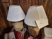 Two matching beige table lamps with white pleated shades on a wooden surface, one lampshade slightly bowed. Both lamps have cords visible, indicating working condition as claimed by seller.