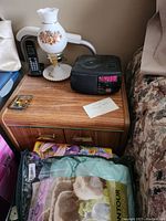 First side table shown topped with a lamp, digital clock radio, and small decorative lamp. Wood grain laminate finish visible on top. Two-door cabinet with brass-colored horizontal handles and trim at base. Wood grain pattern is medium tone.