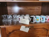 Photo showing five clear etched glass stemware glasses and nine assorted coffee mugs grouped together on a wooden shelf.