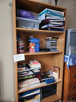 Photo shows a wooden bookshelf filled with various items including binders, plastic containers, CDs, books, and papers. There are three main shelves and one base shelf. The wood finish is brown and the shelf is sturdy.
