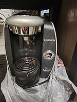 Front and side view of Bosch Tassimo coffee maker showing buttons, cup platform with drip tray, and water reservoir.