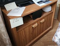 Wooden TV stand showing the front with two cabinet doors and gold handles, open middle shelf, with a TV on top and various papers and remotes.