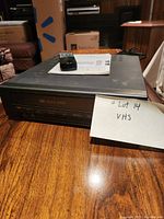 Black VHS player with remote control and manual on top placed on wooden table.