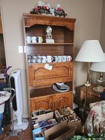 Wood cabinet from front view showing upper shelves holding mugs and glasses (not included), two lower doors, and a drop-down compartment door. Cabinet has medium brown wood finish and classic styling.