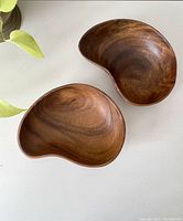 Two vintage wooden kidney-shaped catch all bowls placed on a white surface with a plant visible nearby. Bowls have a smooth finish and natural wood grain.