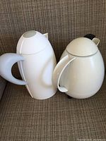 Photo showing two white thermos bottles side by side on a couch upholstery. The Guzzini thermos on left has a matte finish and gray handle; Leifheit thermos on right is rounder with a glossy finish and dome lid.