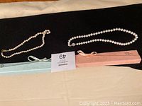Two pearl necklaces laid out flat with their boxes: one necklace with smaller white pearls in a light blue box with bow, and one necklace with larger white pearls in a pink box with bow.
