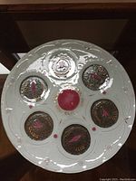 Top view of the round glass seder plate, showing 6 circular depressions with Hebrew text and a central red dot.