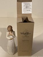 Front view of the Willow Tree Angel of Mine figurine with box showing the figurine holding a child, dressed in white textured dress, standing beside the box.