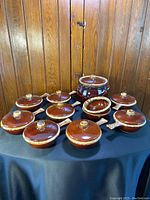 Photo of all 20 brown dripped glaze ceramic soup bowls and one large pot on a black table against wood panel wall.