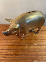 Front angle view of a brass piggy bank on a wooden surface showing the coin slot on top and pig features.