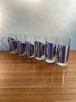 Photo shows six tall clear glass hot toddy glasses with silver steel holders and handles lined up on wooden surface.