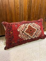 Side view of rectangular red wool Moroccan floor cushion with detailed pattern, placed upright on white textured rug in front of wood paneling.