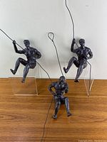 Front view of three ceramic climber sculptures sitting on clear stands with metal wires attached representing climbers hanging on ropes.