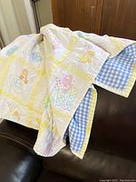 Quilt blanket draped over chair showing pastel Care Bears patchwork and yellow checked border with blue gingham fabric on reverse.