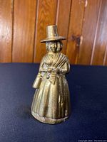Front and angled side view of vintage English brass bell shaped as a Pilgrim woman in traditional attire, positioned against a wooden background.