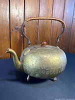 Front angle view of the etched brass teapot showing detail patterns and wicker handle.