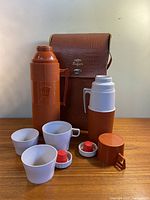 Full set showing two thermoses, four cups (three white plastic, one with handle), two red stoppers and the vinyl carrying case behind.