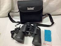 Bushnell binoculars centered with carrying case behind and cleaning cloth to the side on white background.