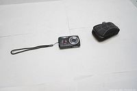 Photo of Kodak digital camera with wrist strap and protective case placed separately on white background.