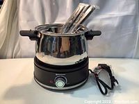 Full assembled fondue set with six wrapped forks inserted in pot, power cord visible