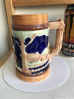 Side view of ceramic beer stein showing relief design with human figures and architectural elements in blue and beige with green background and natural textured handle.