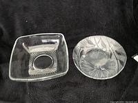 Top view of small square clear glass bowl with circular etched pattern and round translucent glass bowl with sunflower pattern on a black fabric background.