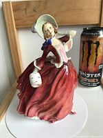 Front view of the Royal Doulton Autumn Breezes porcelain figurine showing the woman in a flowing red dress holding a lantern, with Monster Energy drink can for scale.
