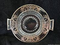 Top view of round glass plate with silver inlay floral and vine patterns around rim and handles, showing design detail and overall condition.