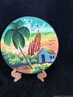 Front view of round clay plate with painted Hawaiian tropical scene, palm tree, flowers, small house and birds at sunset. Signed by artist.