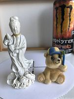 Front view of vintage white porcelain Guanyin figurine standing on lotus flower base alongside a small textured teddy bear figurine holding a blue object.