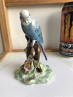 Front view of the blue parakeet perched on brown tree branch with floral base showing detailed painting and texture.